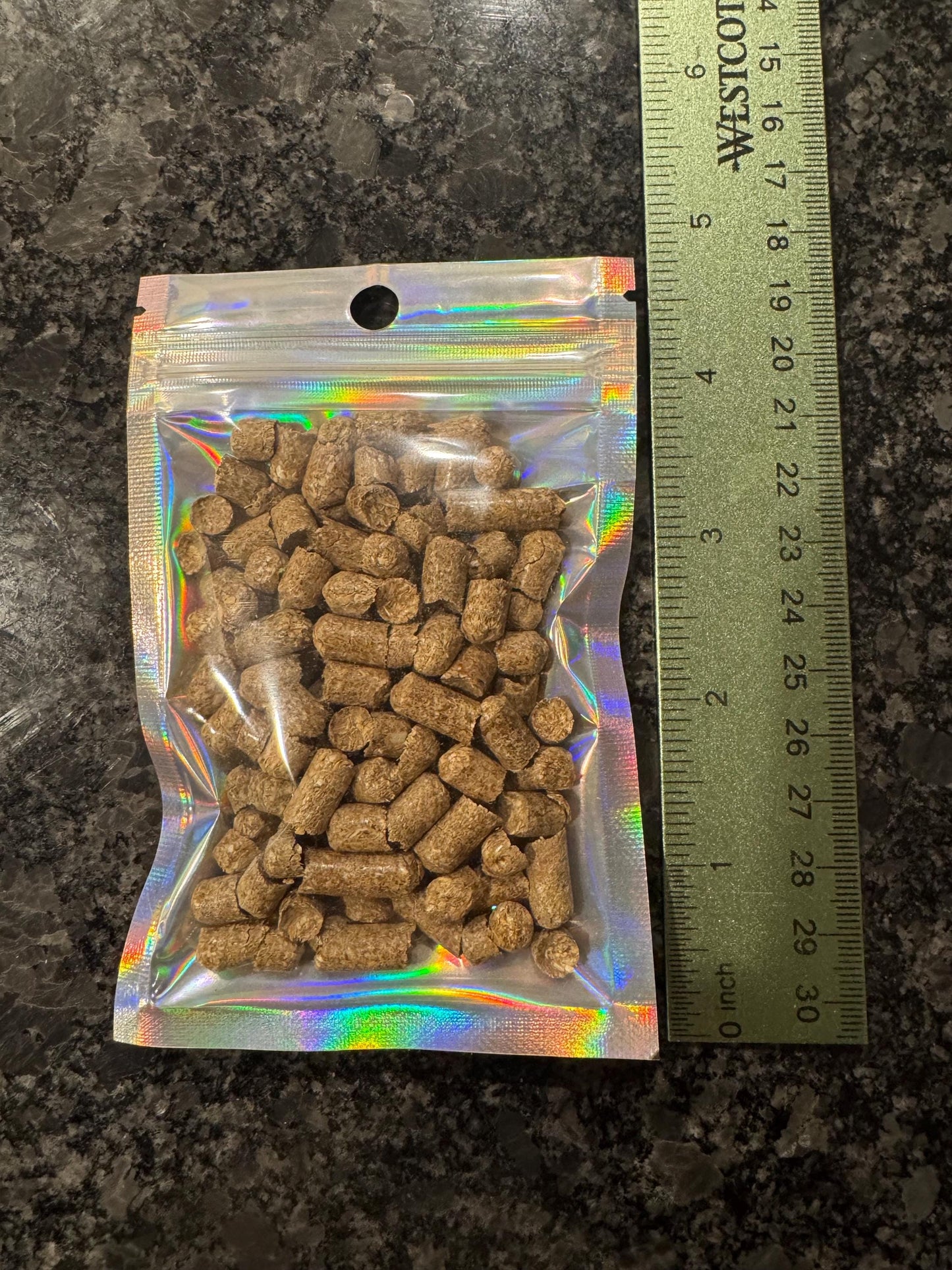 A small, iridescent, resealable bag filled with brown, cylindrical pellets. The bag is next to a ruler showing 4.75 inches long
