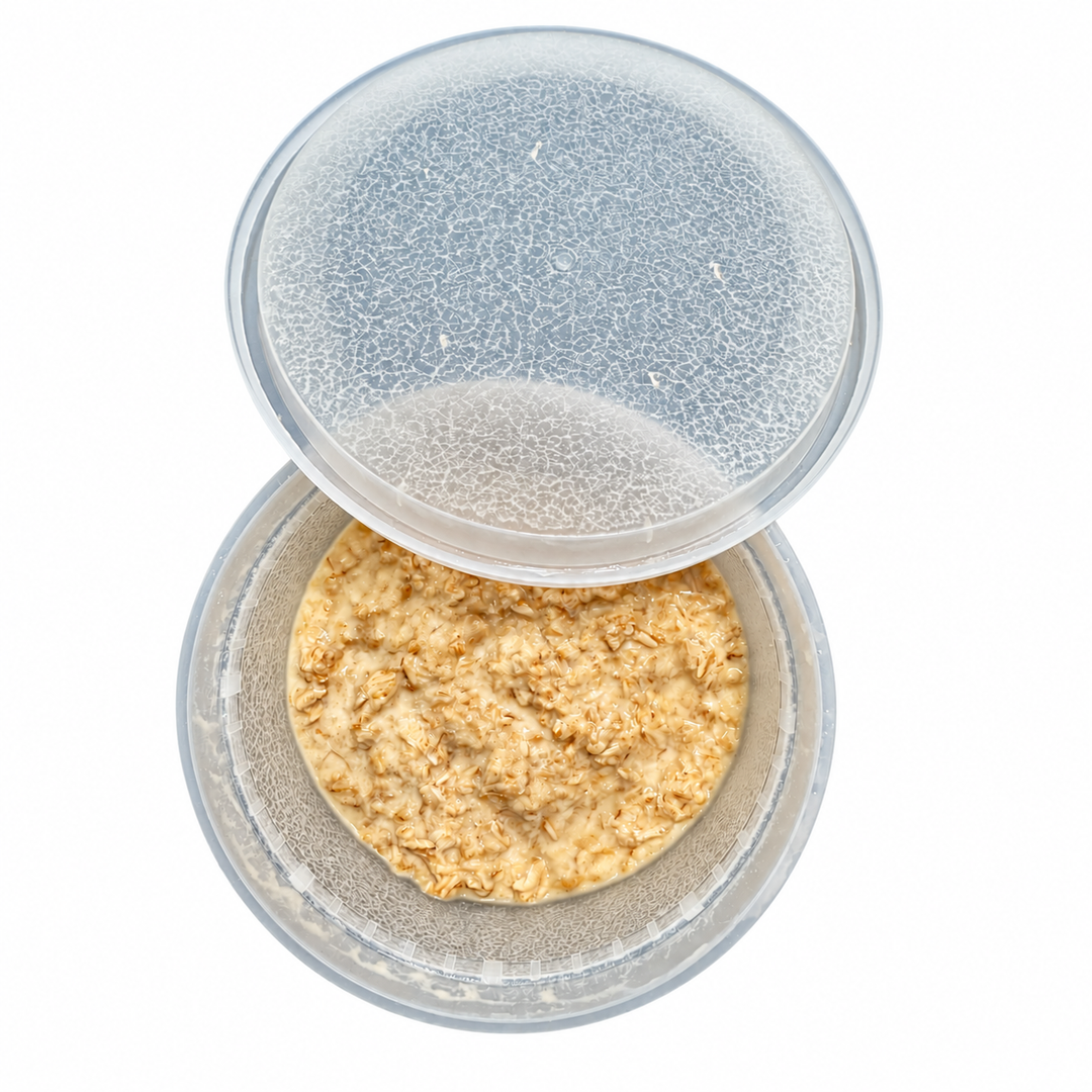 Open microworm culture in a clear deli cup with oatmeal medium and tiny white worms visible on the walls and lid, against a white background.