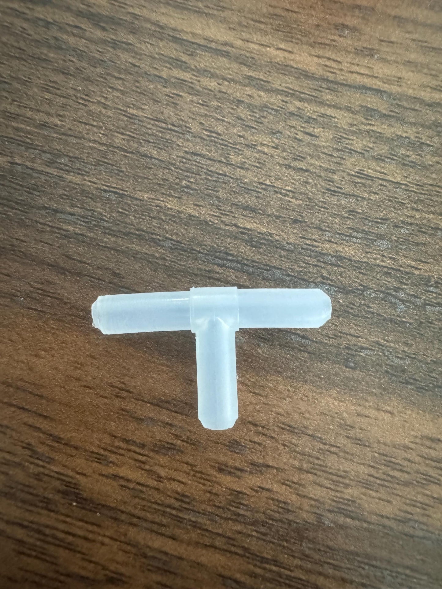 Clear plastic T-shaped aquarium airline tubing connector placed on a wooden surface, showing its three barbed ends for connecting standard 3/16-inch tubing.